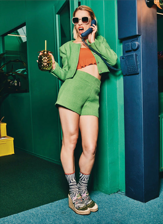 Woman in green outfit talking on a payphone with a drink in hand, standing against a green wall. Xpooos sokkar - bestu sokkarnir - sætir sokkar